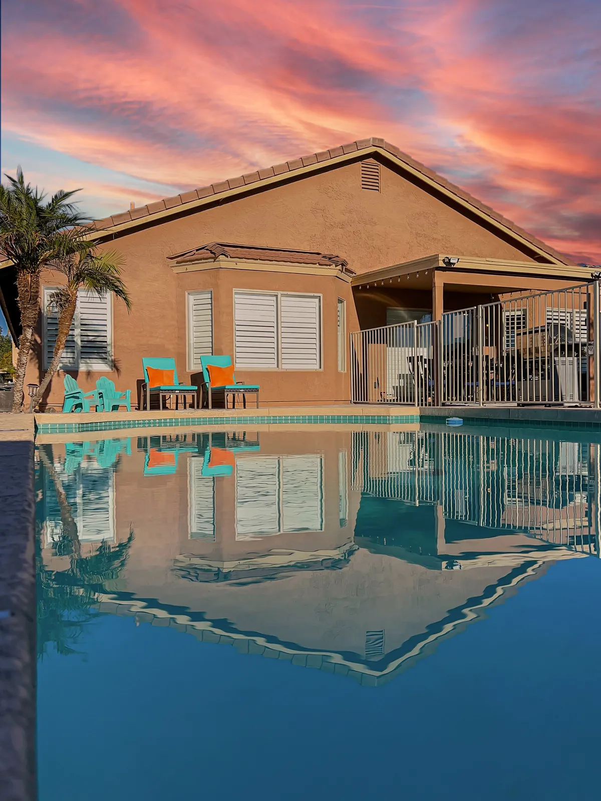 A sparkling pool sits in the backyard of a modern home, surrounded by chairs, a palm tree, and open sky. The scene suggests relaxation and leisure, with plenty of space for groups to enjoy the outdoors together.