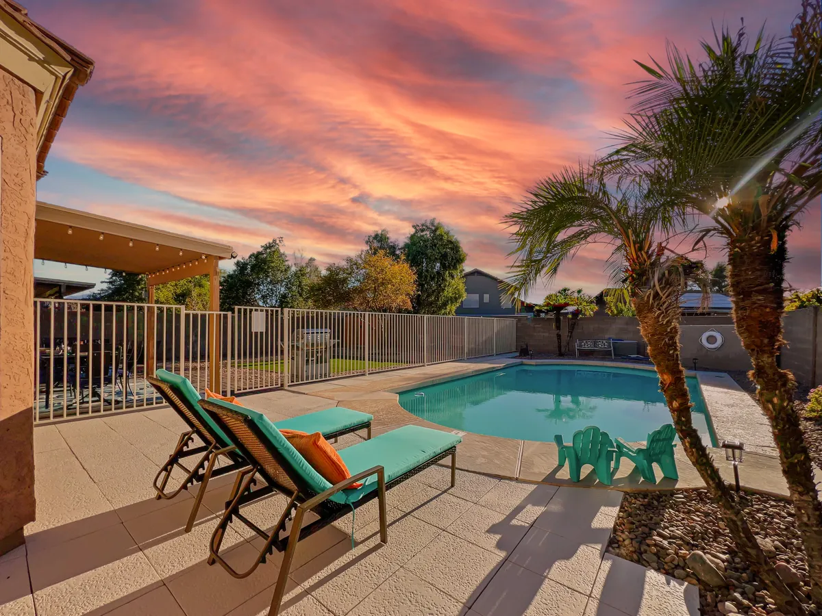 A tranquil backyard scene at sunset featuring a private swimming pool with a safety fence, surrounded by lounge chairs and palm trees. The sky glows with warm colors, creating an inviting atmosphere for evening gatherings and family relaxation.