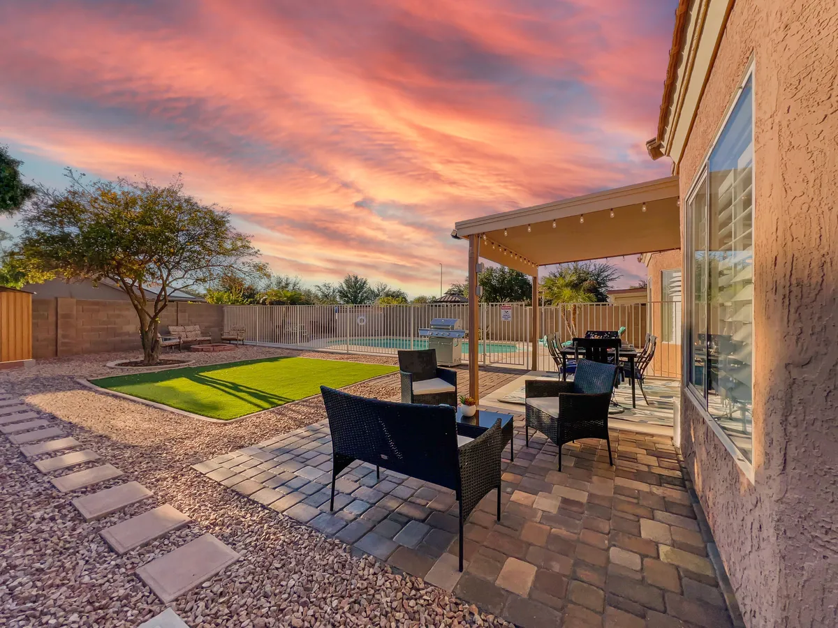 A tranquil patio seating area overlooks the manicured lawn and pool, bathed in the warm glow of a sunset sky. Comfortable outdoor furniture invites guests to relax or dine al fresco, while the pool area promises fun and relaxation.