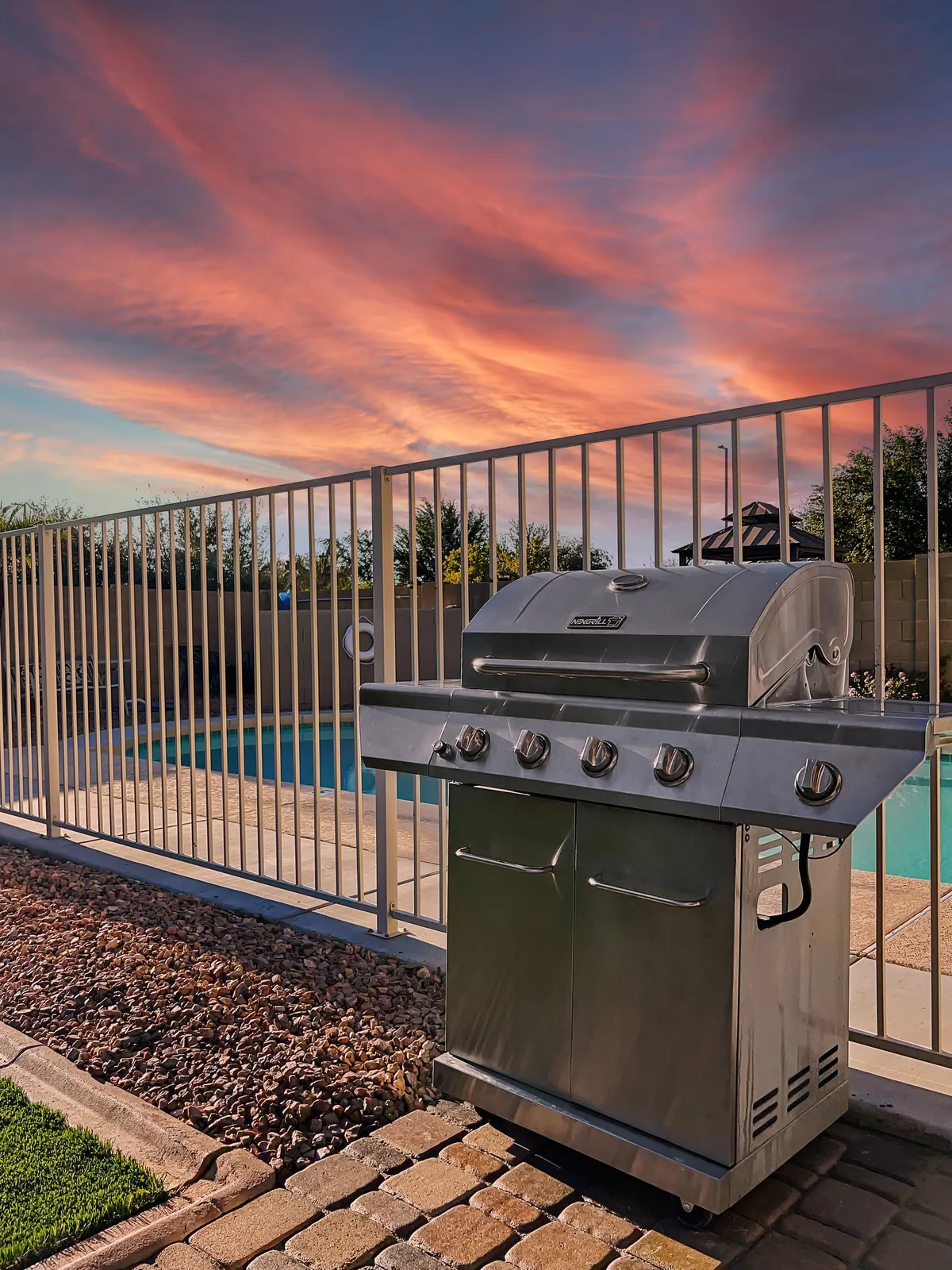 A cheerful backyard scene featuring a grill and a swimming pool set against a stunning sunset sky. The grill is ready for barbecuing, while the pool area invites guests to gather, swim, or relax during lively outdoor events.