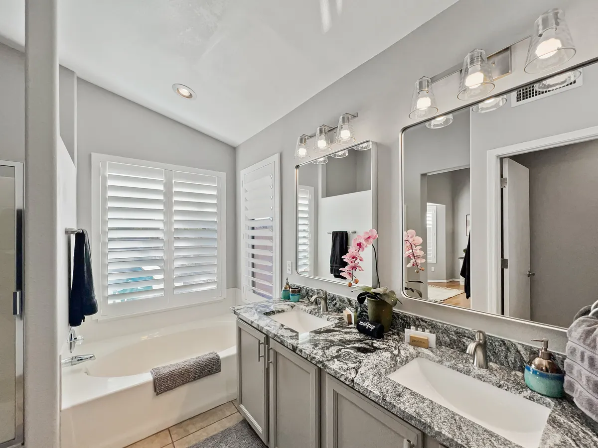 A serene spa-like bathroom with a double sink vanity, bathtub, mirrors with lights, window shutters, and decorative plants. The space exudes calm and luxury, perfect for unwinding with a relaxing bath or preparing for the day ahead.