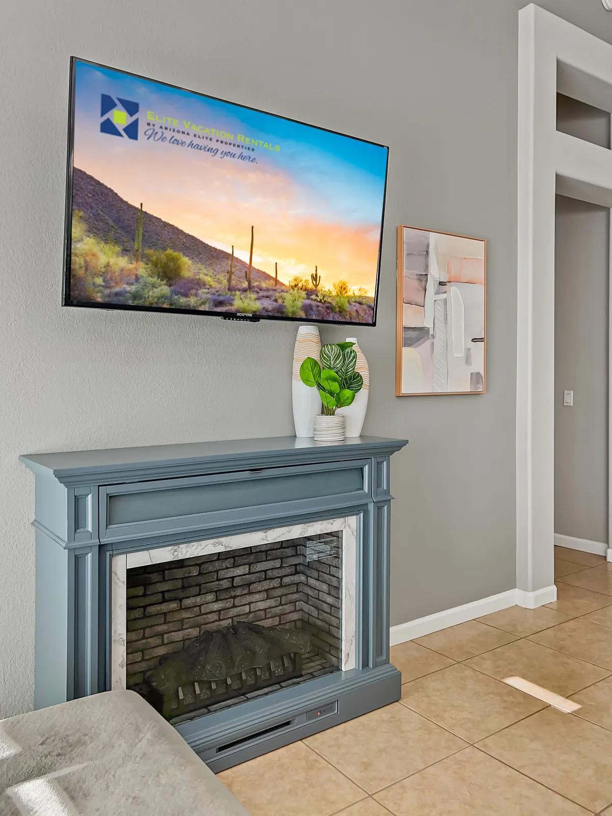 A cozy living area with a fireplace, TV, modern artwork, and greenery. The space is styled for relaxation and communal enjoyment, with focal features that invite gatherings around both the fire and television.