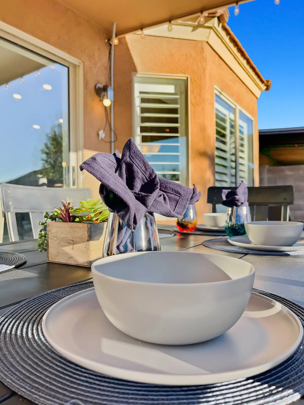 An inviting outdoor dining area set with a stylish table arrangement, succulent centerpiece, and dinnerware. The scene hints at shared meals, laughter, and late-night recaps of the day's baseball highlights, all against a soft, sunset-tinged sky.
