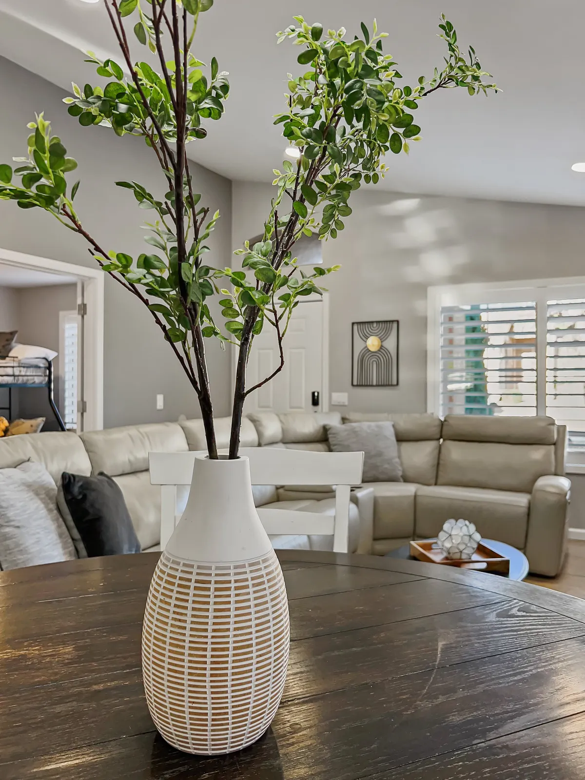 A bright, airy living room with a light-colored sectional sofa, a dark wooden table, and a striking decorative vase. The arrangement is conducive to intimate conversations and relaxed entertaining, reflecting a space ideal for connecting with friends and family.
